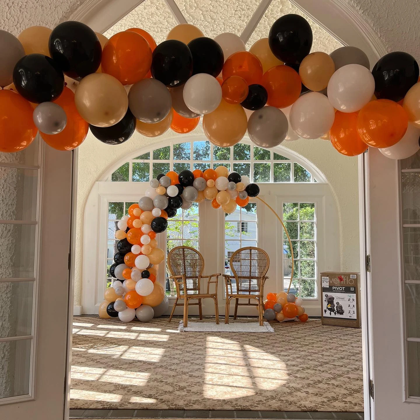 Modern 'you are my sunshine' baby shower photo station with an orange, black, and grey balloon arch over two wicker chairs.