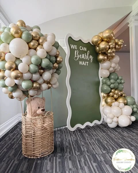 A teddy bear sits inside a wicker basket with a large bunch of sage green, white, and gold balloons attached above it to look like a hot air balloon.