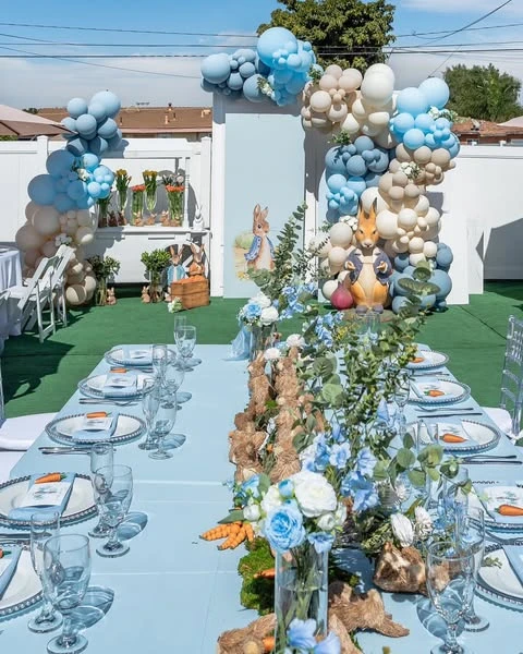 A long table set for a Peter Rabbit baby shower with a lush centerpiece of rabbit figurines, carrots, and blue flowers.