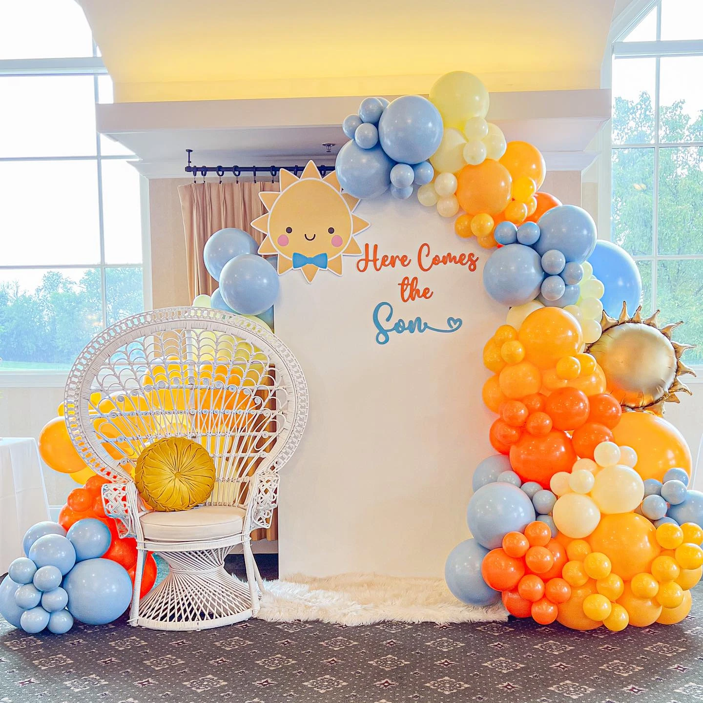 Here Comes the Son baby shower photo backdrop with a colorful balloon arch and a white peacock chair.