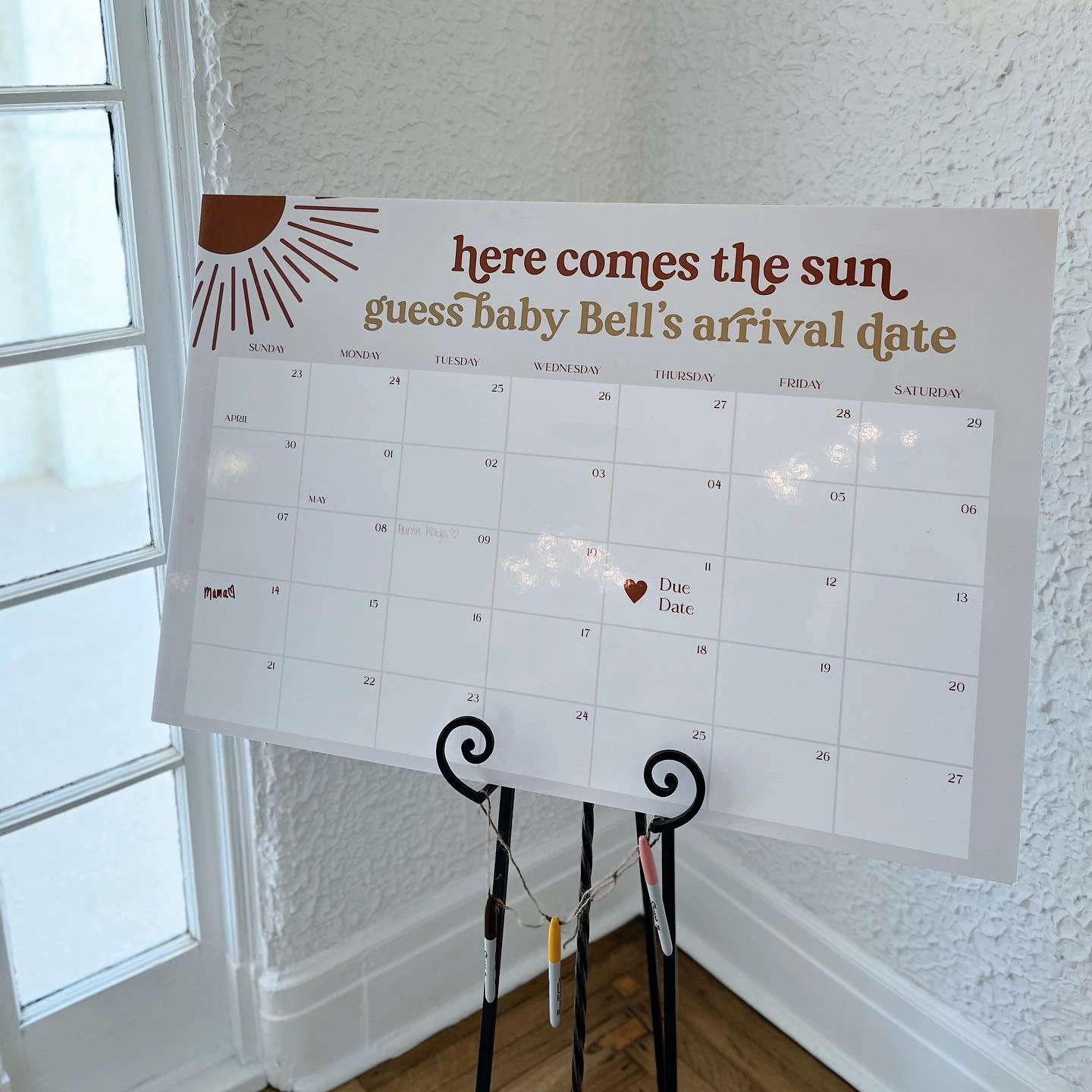 A 'here comes the sun' baby arrival date guessing game on a calendar poster.