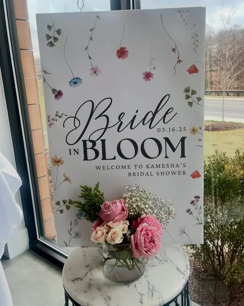 A 'Bride in Bloom' welcome sign for a spring bridal shower, decorated with watercolor wildflowers and a vase of fresh pink roses.