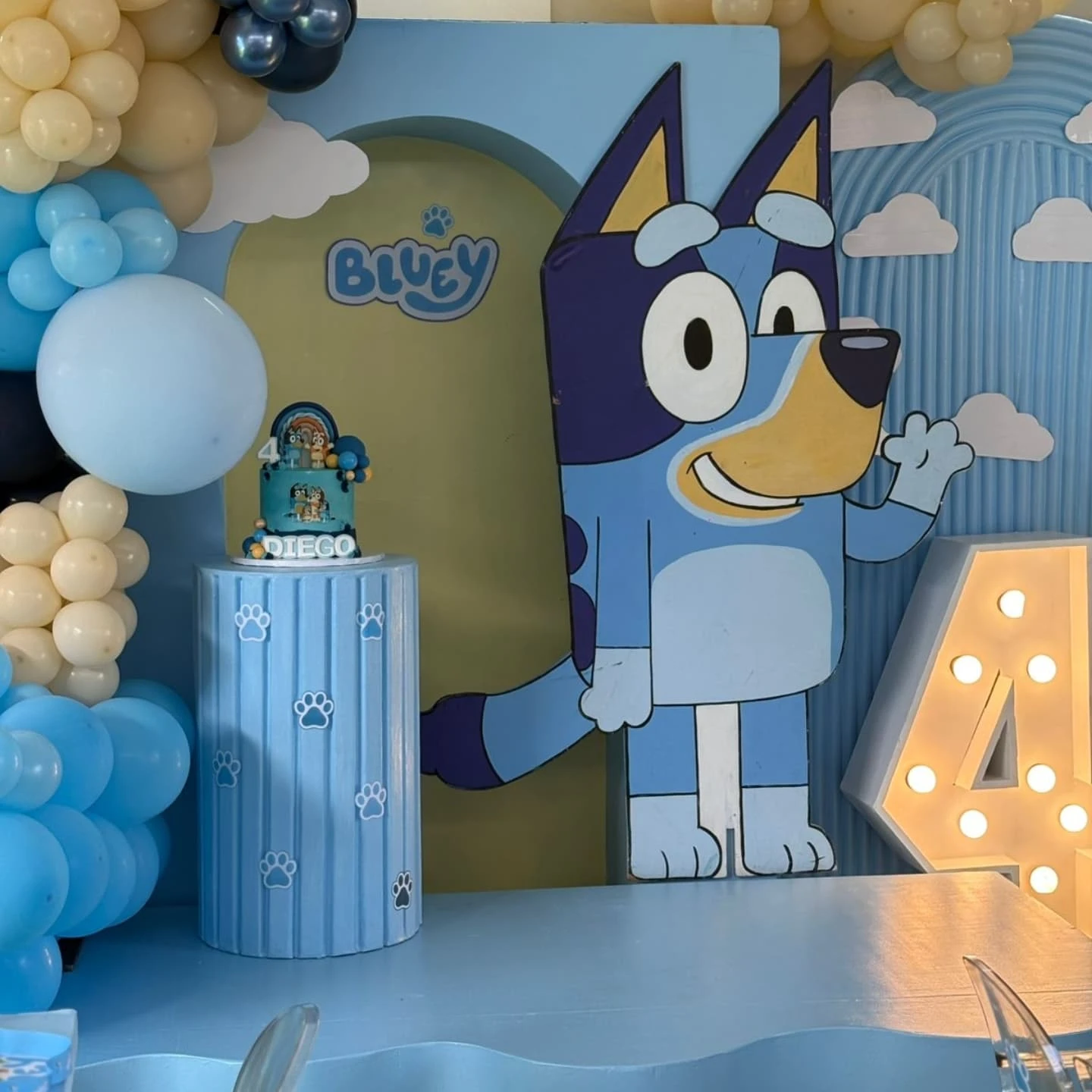 A large Bluey character cutout stands next to a light-up number 4, a themed birthday cake on a blue pedestal, and a multi-toned blue and yellow balloon garland.