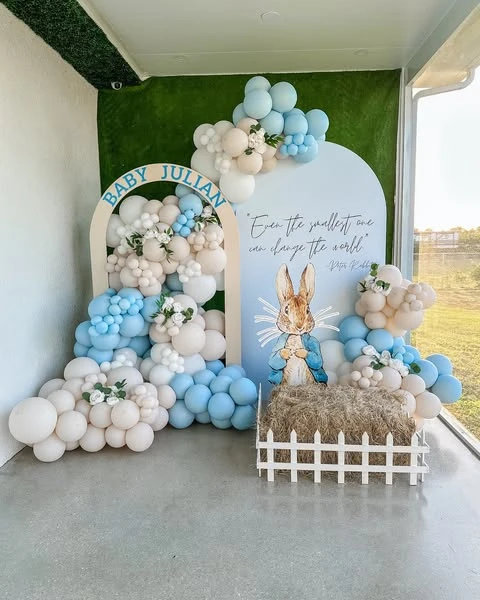 A Peter Rabbit baby shower photo backdrop with a large character cutout, a hay bale, and a blue and white balloon garland.