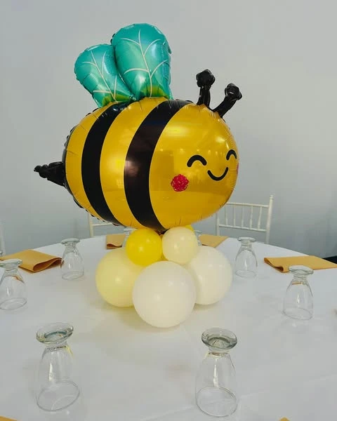 A baby shower centerpiece with a large, smiling bumblebee Mylar balloon floating above a cluster of white and yellow latex balloons on a table.