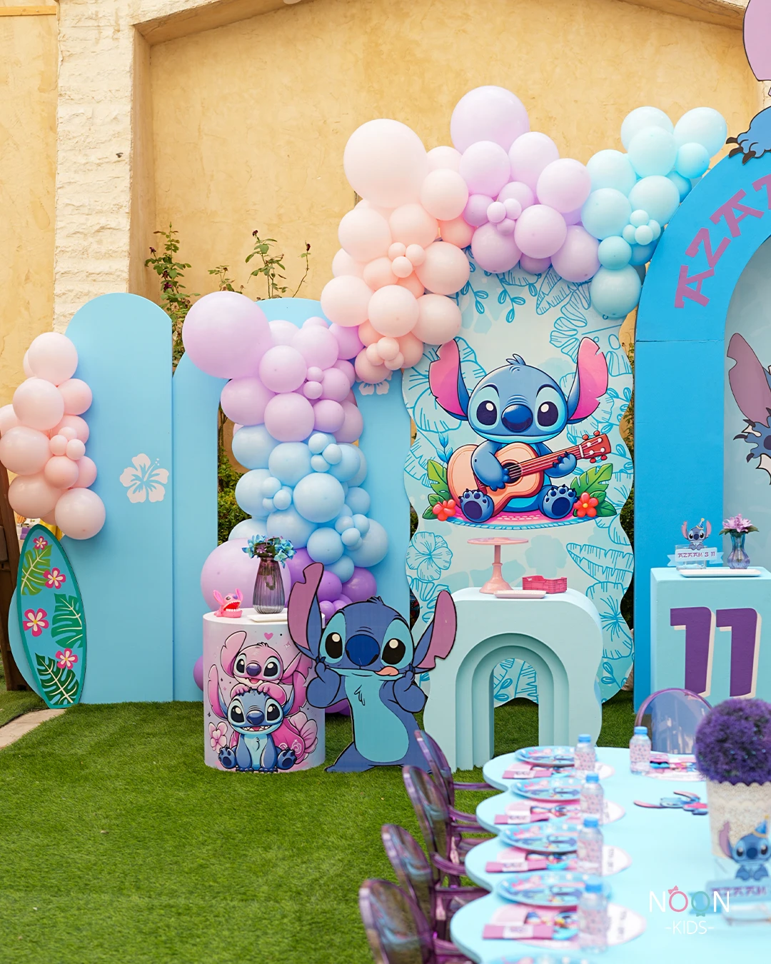 Large character cutouts of Stitch and Angel stand in front of a pastel balloon garland at a birthday party.