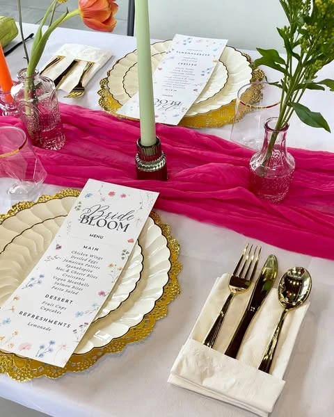 A spring bridal shower place setting with a floral menu, gold cutlery, and a bright pink table runner.