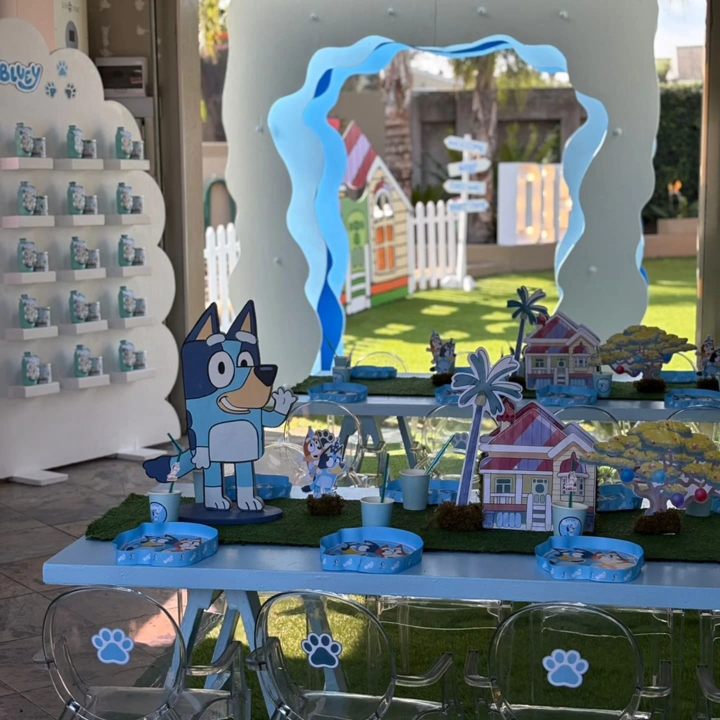 A Bluey-themed kids' party table with a small Bluey cutout, looking towards a white, cloud-shaped party favor display shelf in the background.