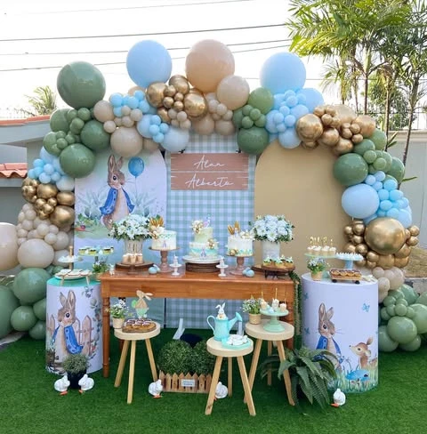 A Peter Rabbit themed baby shower dessert table with a large, multi-colored balloon arch over a layered gingham and character backdrop.