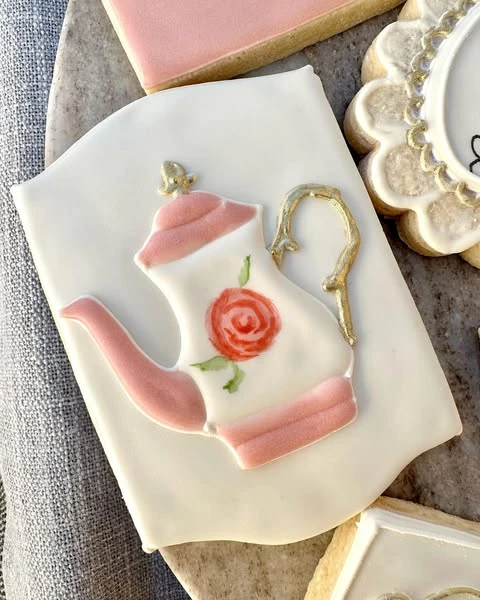 A detailed teapot-shaped cookie with pink accents and a painted rose.