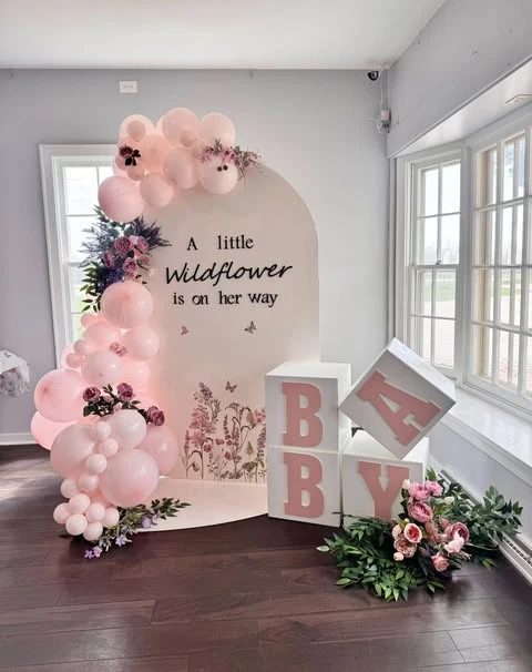A wildflower baby shower backdrop with the text 'A little wildflower is on her way' framed by a pink balloon garland and floral arrangements.