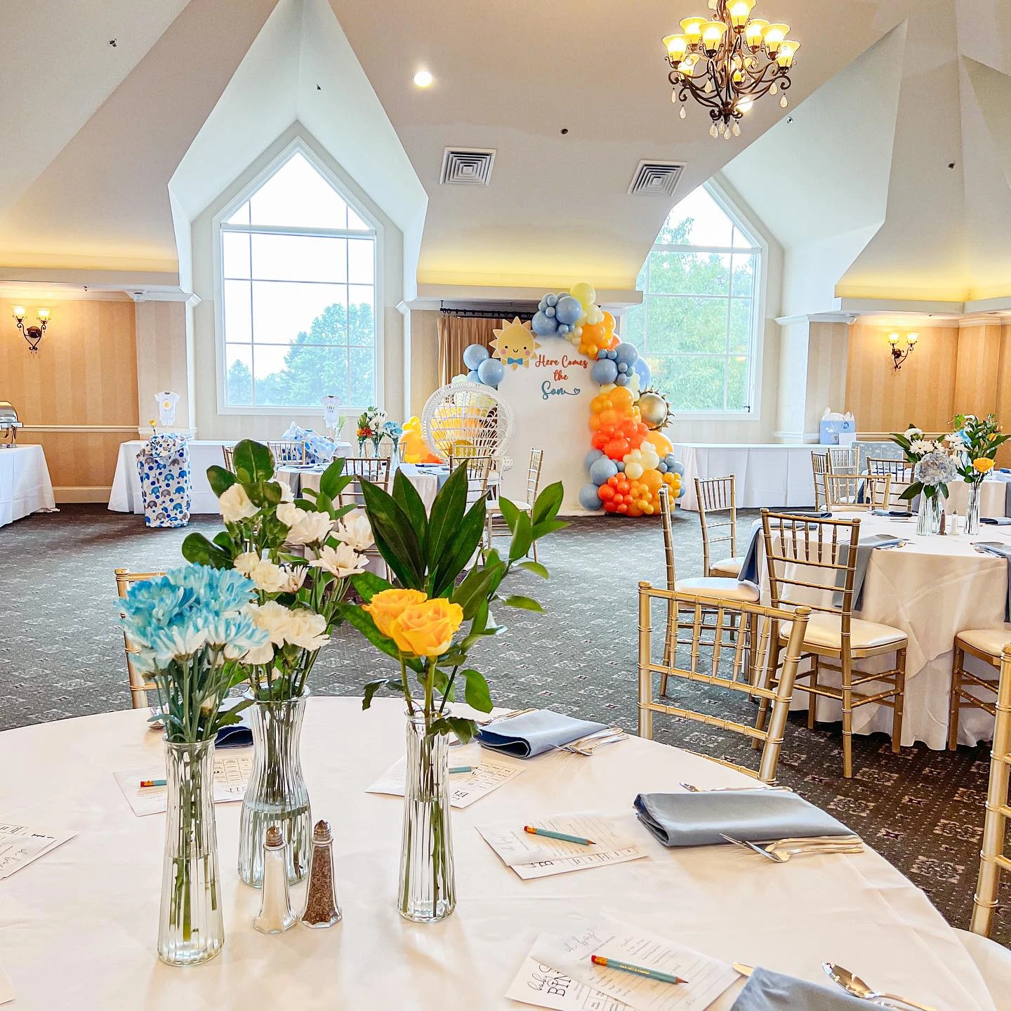 Wide view of a baby shower event space, showing the 'Here Comes the Son' balloon backdrop as the main focal point in the room.