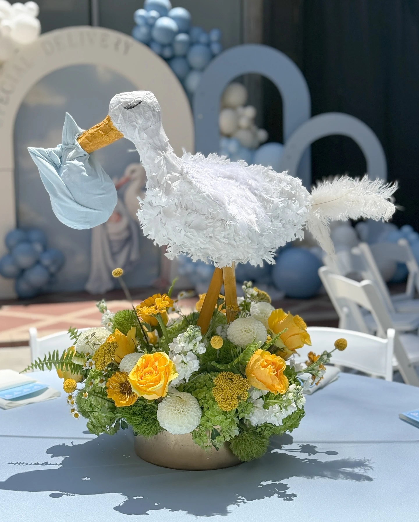A baby shower centerpiece featuring a white paper-mache stork figure holding a blue bundle, standing in a gold bowl filled with yellow and white flowers.