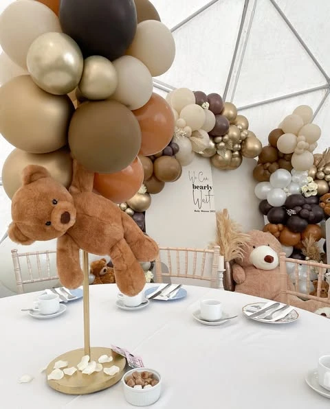 Teddy bear centerpiece on a gold stand for a 'We Can Bearly Wait' baby shower, surrounded by brown, tan, and gold balloons.