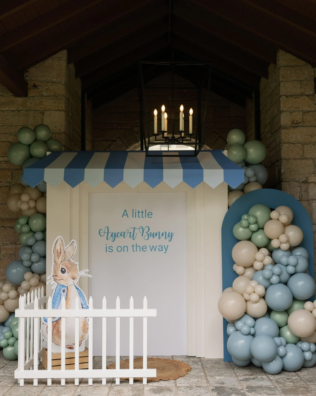 Peter Rabbit baby shower photo backdrop with a character cutout, picket fence, and a blue and green balloon arch.