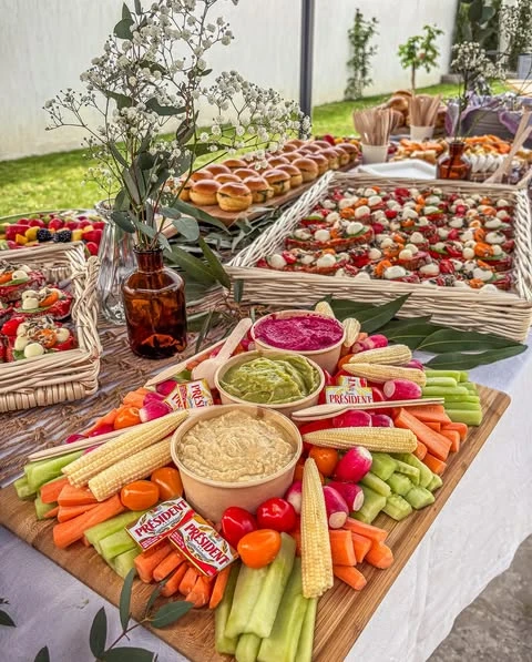 A large wooden board arranged as a crudités platter with carrots, celery, radishes, and three bowls of different colored dips for a baby shower.