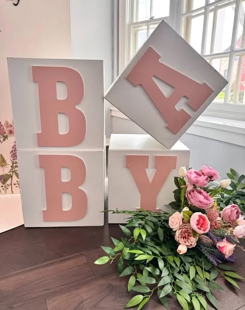 A lush floral arrangement with pink and cream flowers and abundant green foliage, placed on a dark wood floor for a baby shower.