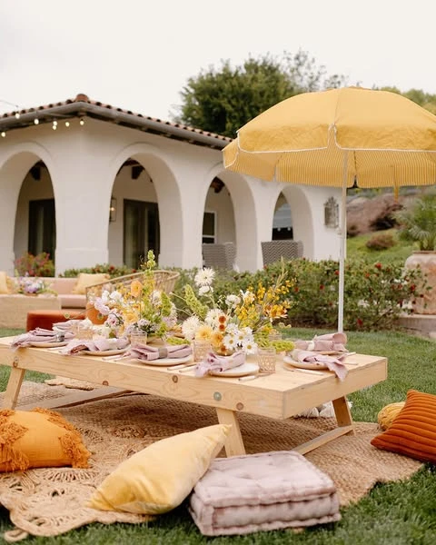 Outdoor bohemian baby shower picnic with a low wooden table, floor cushions, and a yellow umbrella.