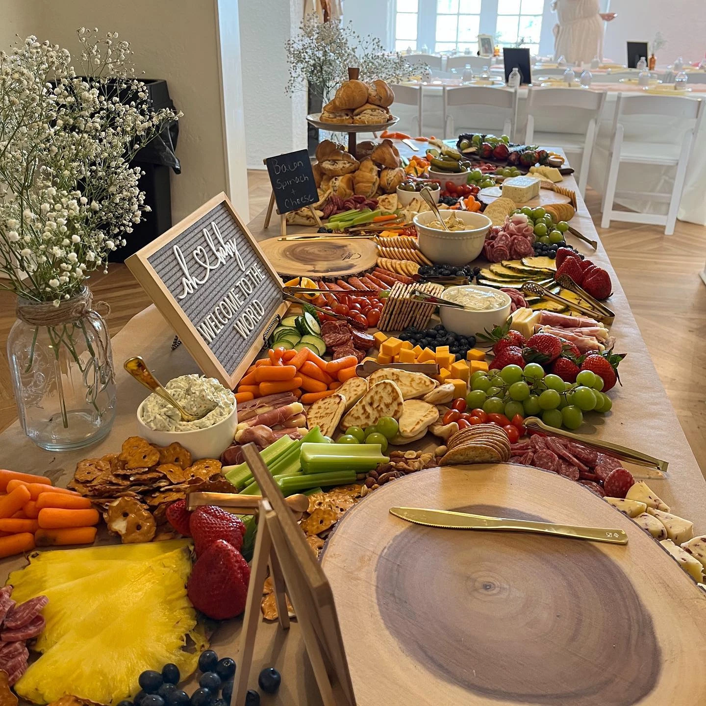 A long grazing table at a boho baby shower, featuring fresh fruits, cheeses, meats, and a letterboard sign.