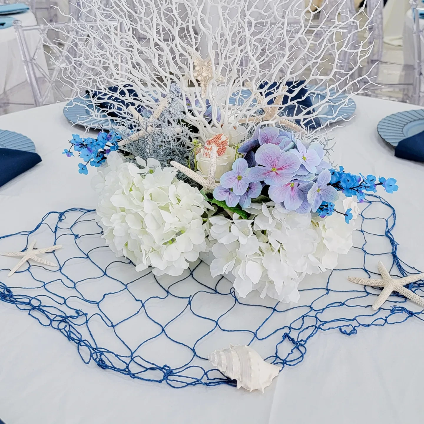 An elegant nautical baby shower centerpiece with a large white fan coral rising behind blue and white hydrangeas on a blue fishing net.