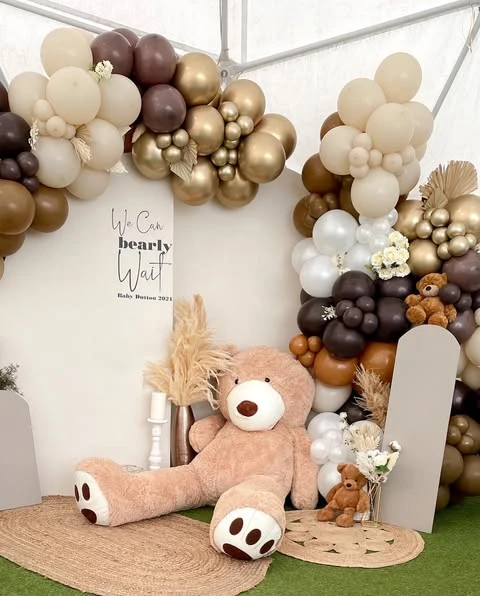 Baby shower backdrop with a 'We Can Bearly Wait' sign, a neutral balloon arch, and a large plush teddy bear on a woven mat.