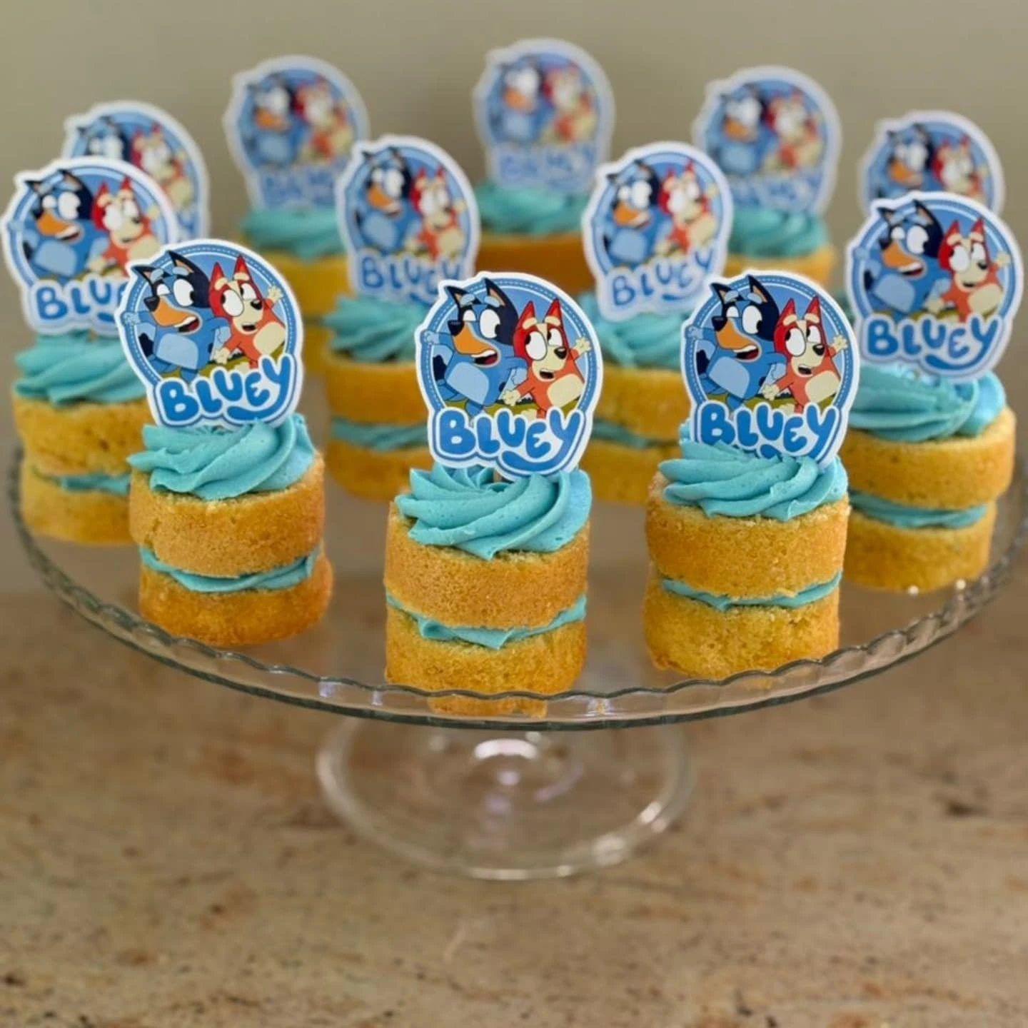 Glass cake stand with individual mini cakes topped with blue frosting and Bluey and Bingo paper toppers.