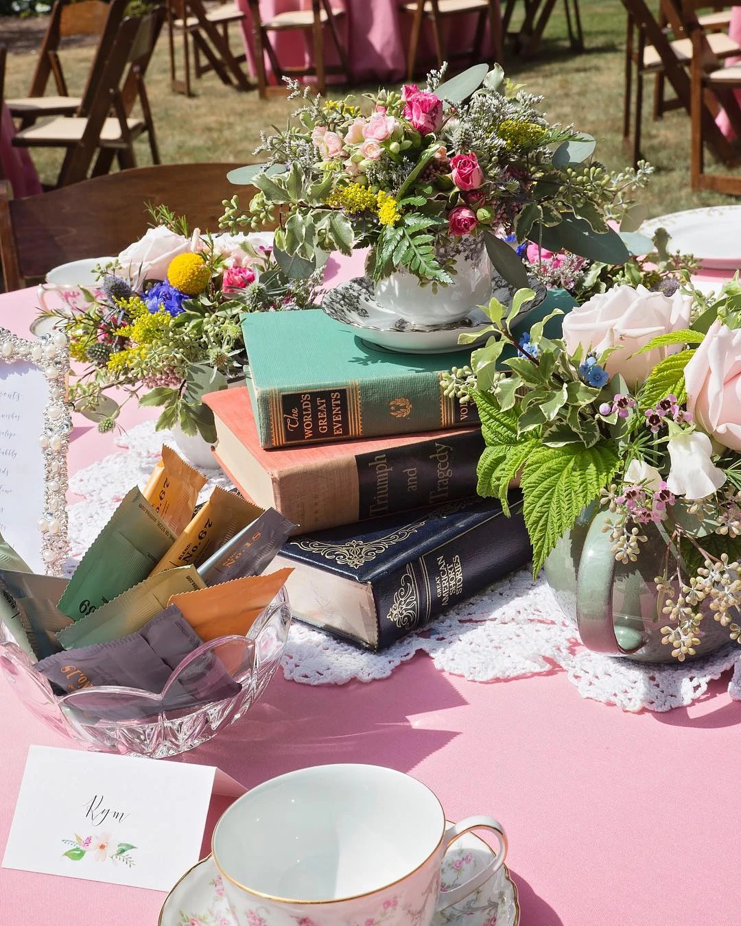 A tea party bridal shower centerpiece with vintage books, flowers in teacups, and a bowl of tea bags on a pink tablecloth.