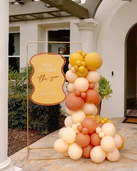 A 'Here Comes the Sun' baby shower welcome sign with a modern balloon garland in yellow and terracotta.