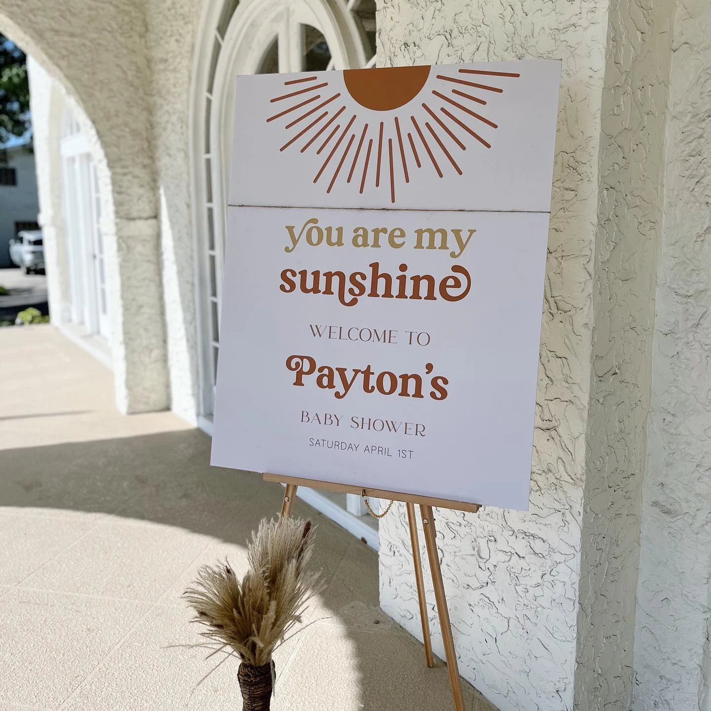 A 'You are my sunshine' baby shower welcome sign on a gold easel with a boho sun graphic.