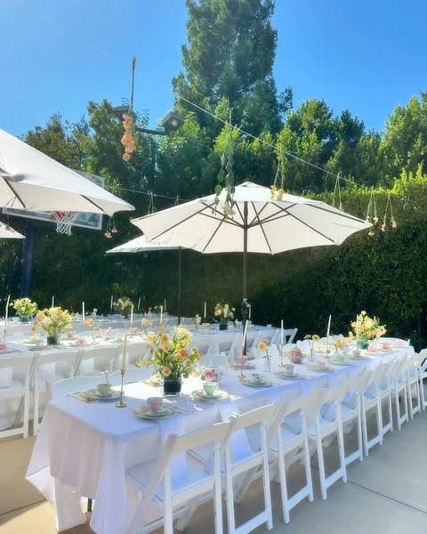 Long white tables set for an outdoor tea party bridal shower under large umbrellas.