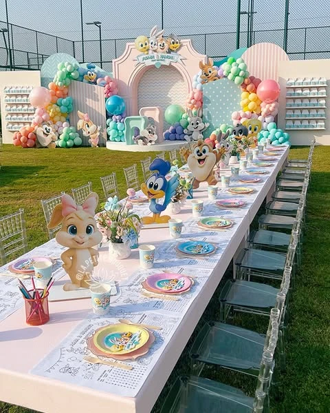 A long baby shower guest table featuring large, freestanding Baby Looney Tunes character cutouts alternating with small flower vases as centerpieces.