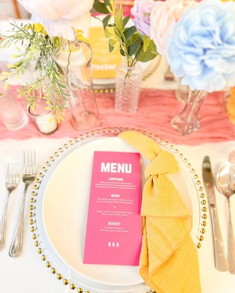 A spring bridal shower place setting with a gold beaded charger, a bright pink menu, and a yellow knotted napkin.