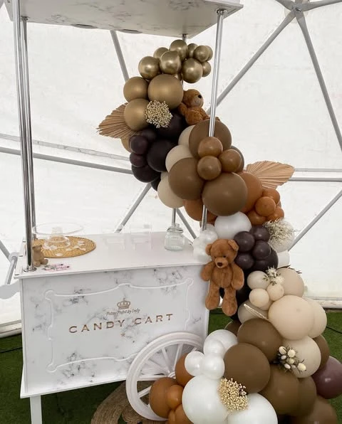 White candy cart decorated with a neutral balloon garland and small teddy bears for a baby shower.