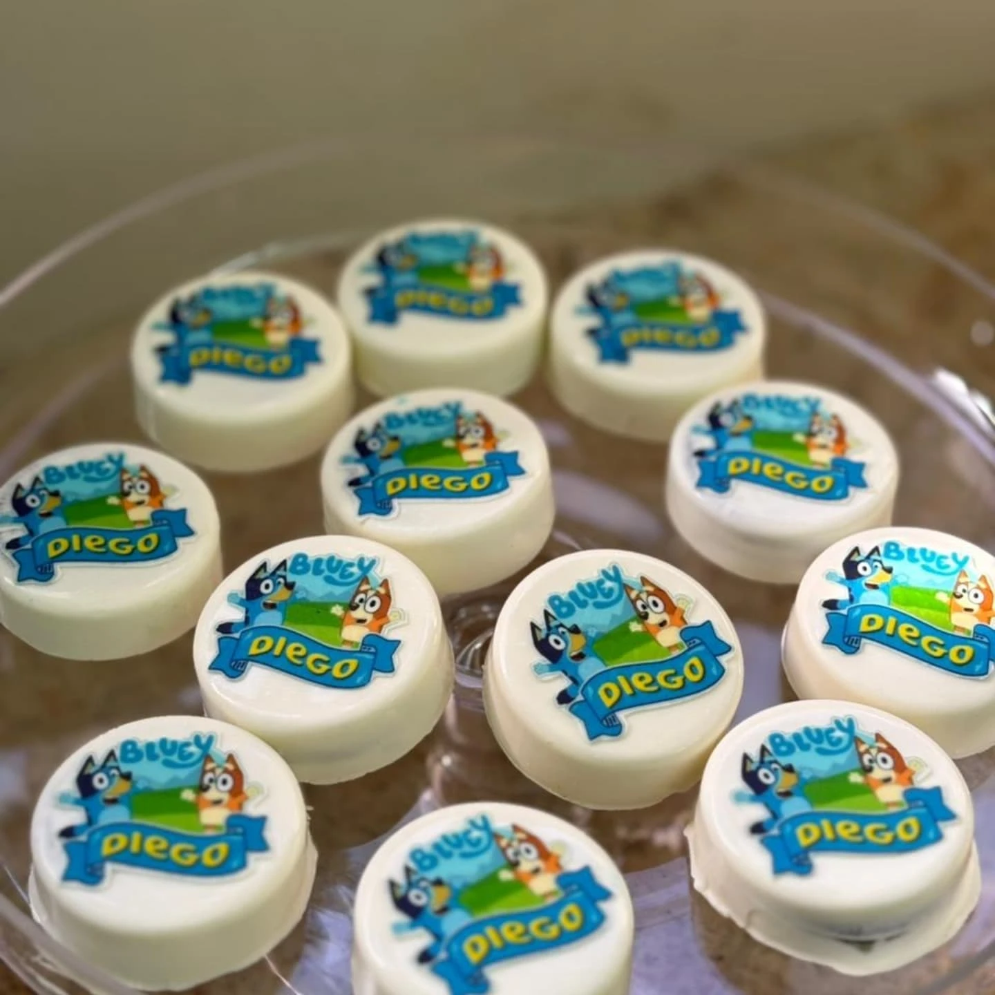 A clear glass platter holds white chocolate-covered cookies, each topped with a colorful edible image of Bluey and Bingo.