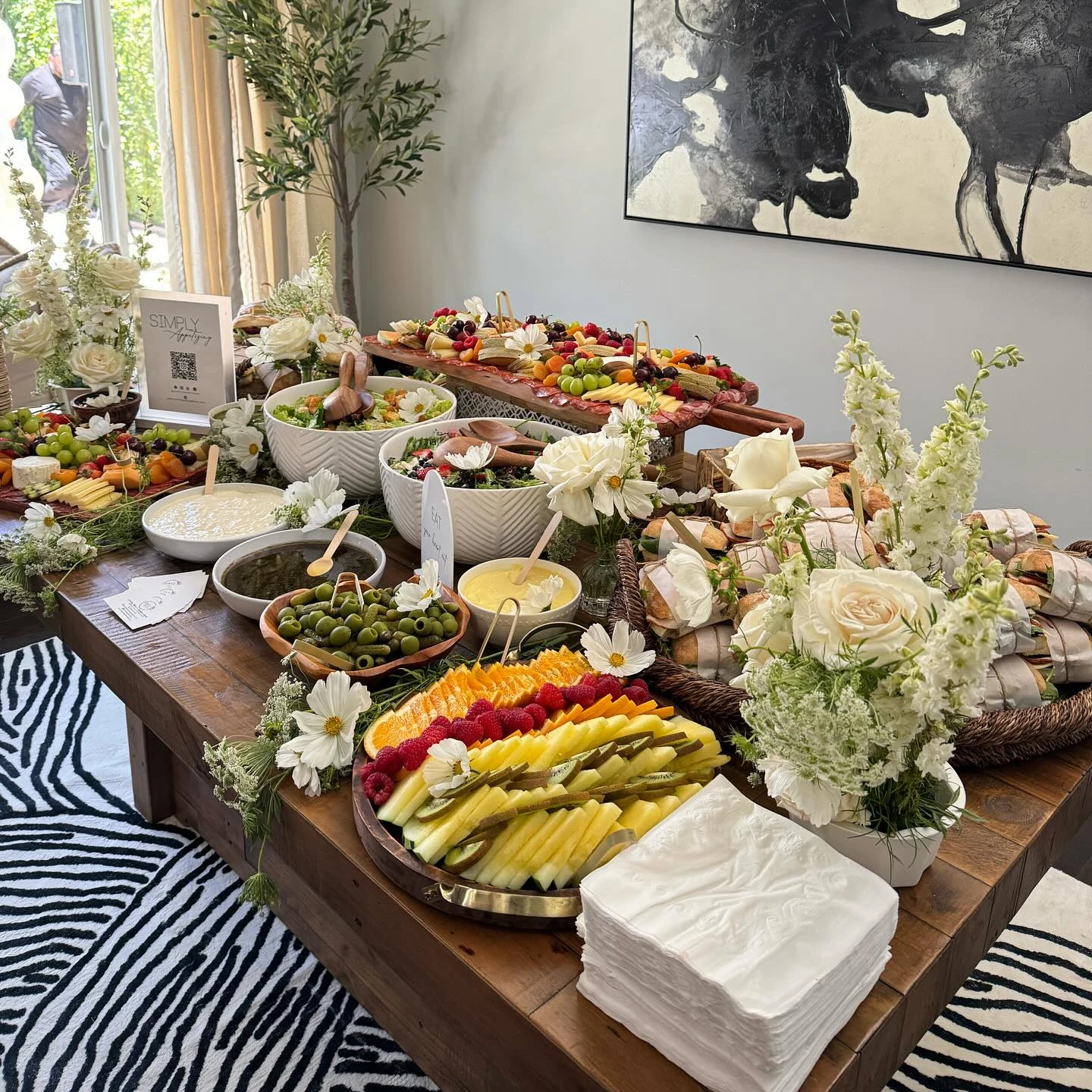 An angled view of a baby shower grazing table, showing wrapped sandwiches, a platter of sliced fruit, and bowls of olives and dips.