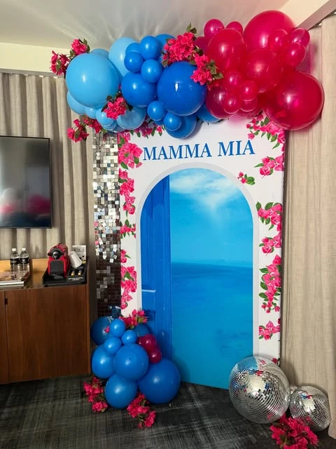 A Mamma Mia bachelorette party photo backdrop featuring a Greek blue door and a vibrant balloon garland.
