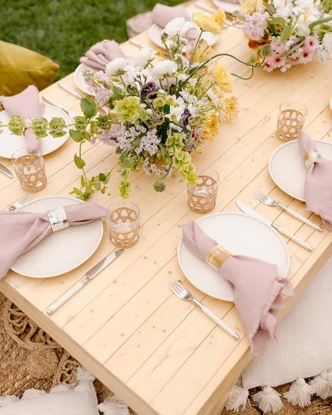 A of a bohemian picnic tablescape with dusty rose linen napkins, woven glasses, and white plates on a light wood table.