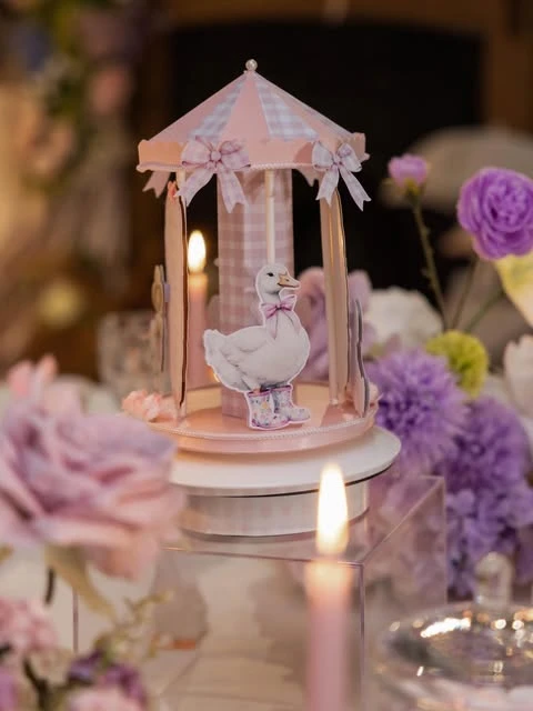 A pink gingham papercraft carousel with a goose illustration, set on a clear acrylic block as a baby shower centerpiece, surrounded by flowers and candles.