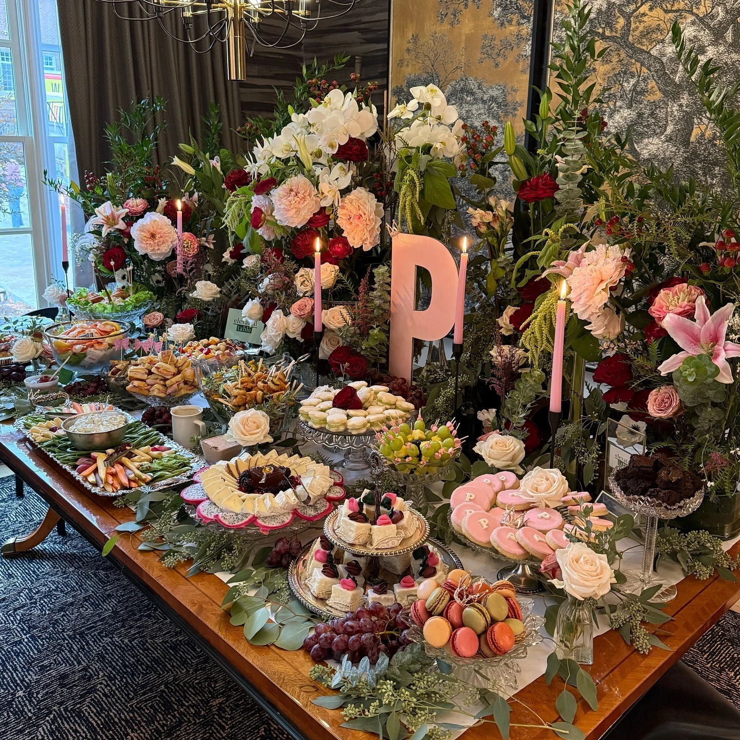 Lush baby shower food table with large floral arrangements, tiered platters of food, and pink candles.