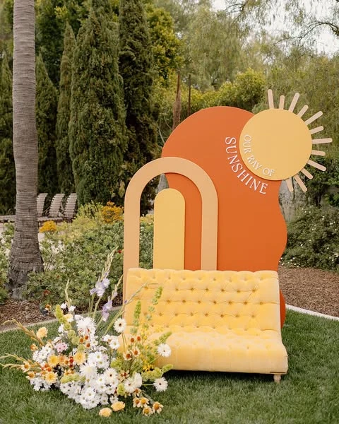 A 'Our Ray of Sunshine' photo backdrop with layered arches and a yellow tufted bench for a baby shower.