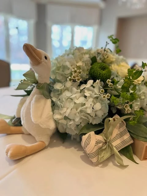 A baby shower centerpiece on a white tablecloth, featuring a cream plush goose toy nestled next to a floral arrangement of green hydrangeas and a small, ribbon-tied baby onesie.