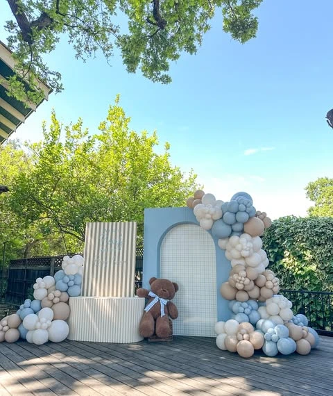 A 'we can barely wait' baby shower backdrop with a blue and brown balloon garland and a large teddy bear.
