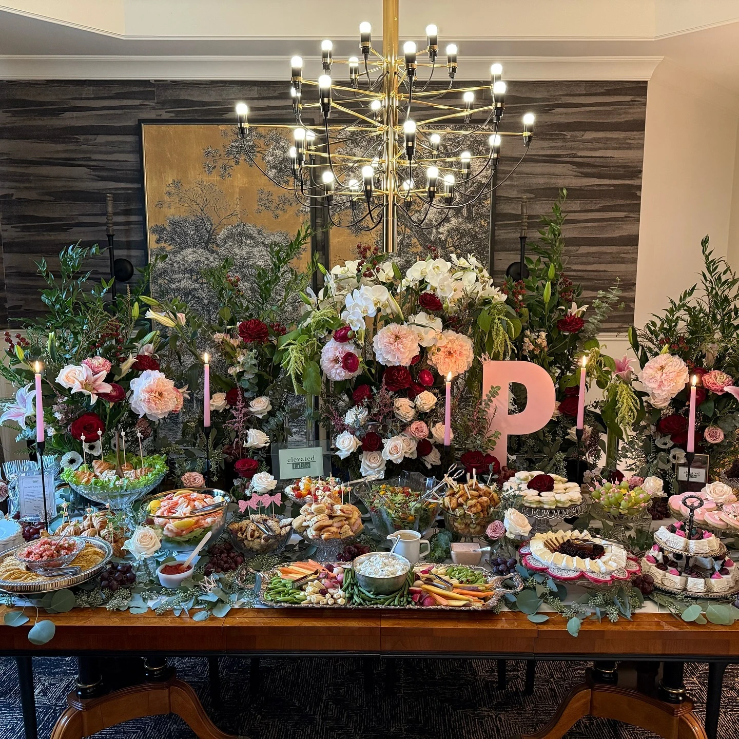 Wide view of a baby shower food table with large floral arrangements, showing savory foods like shrimp cocktail on one side and desserts on the other.