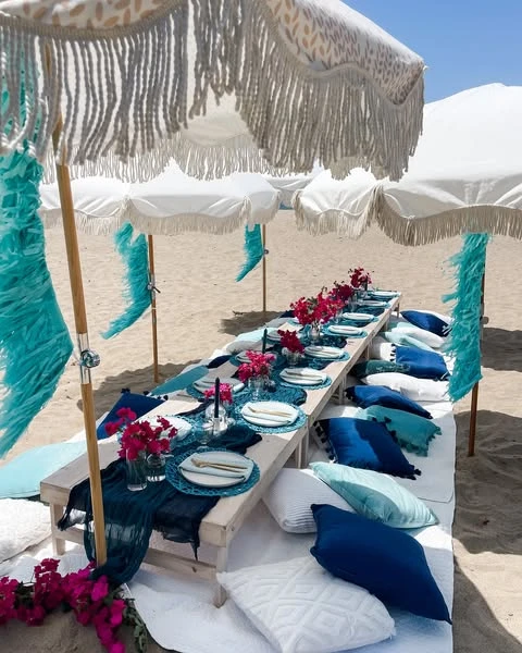 Long low picnic table on a sandy beach under fringed umbrellas for a Mamma Mia themed bachelorette party.