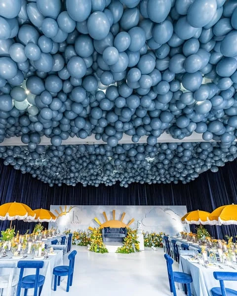 Wide view of a 'Here Comes the Sun' baby shower with a massive blue balloon ceiling, tables set with yellow umbrellas, and a sun-themed backdrop.
