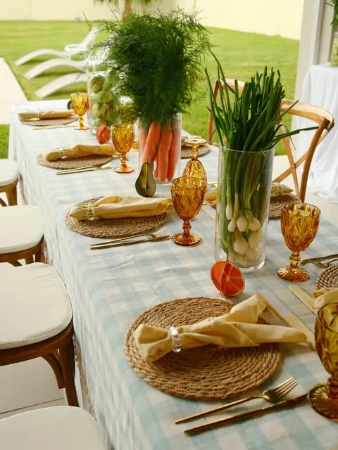 A baby shower table centerpiece made from tall glass vases filled with fresh carrots and spring onions on a blue gingham tablecloth.