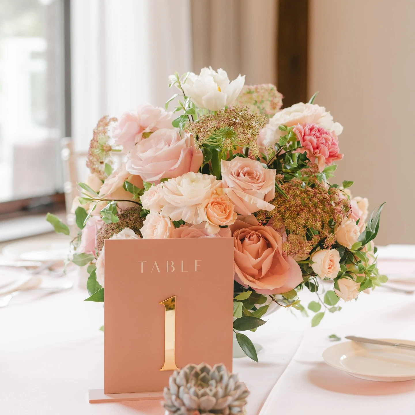 A spring floral centerpiece with pink and cream roses next to a dusty rose table number with a gold numeral.
