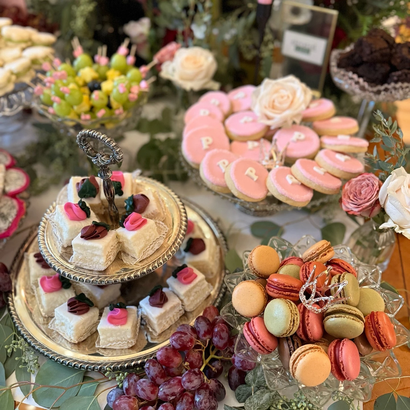 A of a baby shower dessert table featuring a silver two-tiered stand with petit fours, a glass dish of colorful macarons, and a platter of pink monogrammed cookies, all surrounded