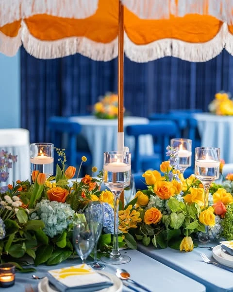 of a baby shower tablescape featuring a yellow umbrella, a floral centerpiece with yellow and orange flowers, and floating candles.