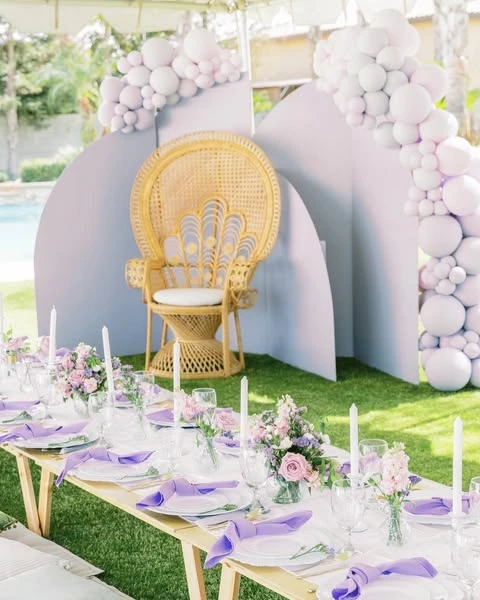 Outdoor spring bridal shower with a lavender and pink theme, featuring a low picnic table, a peacock chair, and a balloon arch.